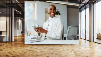 Cheerful young businesswoman working with a smartphone and a laptop at her office desk Wall mural