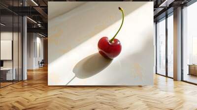 A single fresh cherry with a green stem sitting on a pristine white surface highlighted by natural light Wall mural