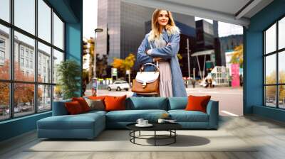 Outdoor fashion lifestyle portrait of blonde pretty young businesswoman, walking at modern buildings area, wearing blue coat and feminine grey dress. Wall mural