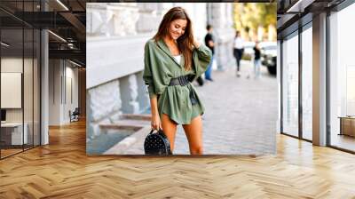 Trendy street style image of amazing fashionable young pretty woman posing on the street, wearing mini shirt dress and small backpack, warm colors, horizontal, sunny summer time. Wall mural