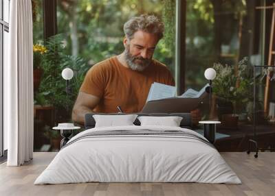 A bearded man in a casual orange shirt reviews documents at a wooden desk, with a green and serene background, illustrating focus and a love for nature. Wall mural