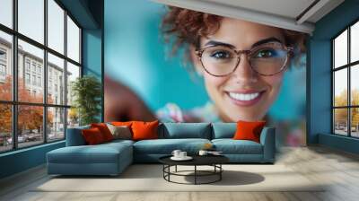 A cheerful woman with curly hair and stylish glasses, smiling and pointing towards the camera, set against a blurred blue background for a lively effect. Wall mural