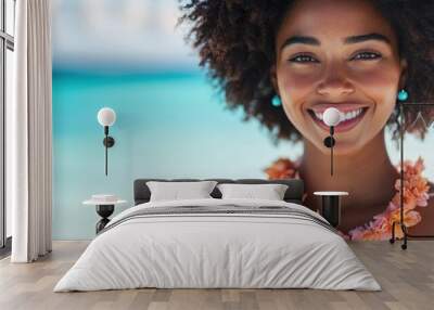 A happy woman with an afro hairstyle wears a flower necklace and smiles brightly against a blurred backdrop of a turquoise ocean on a sunny day. Wall mural