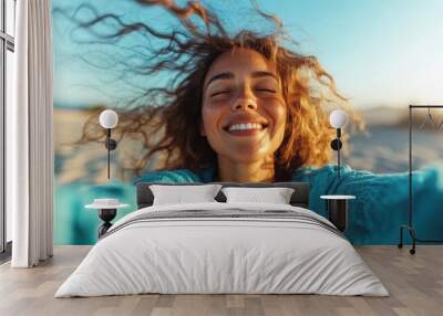 A joyful woman with curly hair closes her eyes and smiles broadly in a sunny beach setting, embodying freedom and happiness in a vibrant blue sweater. Wall mural