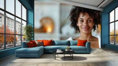 A joyful woman with natural curly hair and soft skin, smiling warmly in a cozy spa environment with soothing background elements and warm lighting. Wall mural