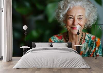 An elderly woman with beautiful curly white hair smiles warmly while wearing a floral top, exuding elegance and positivity against a natural, leafy background. Wall mural