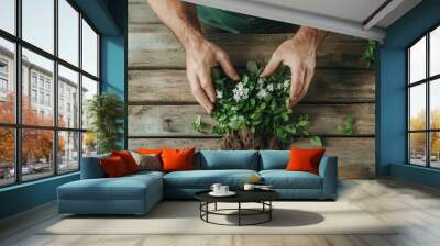 Two hands gently hold a small herb plant with blossoms, representing nurturing and care for nature, surrounded by a rustic wooden table in a calm setting. Wall mural