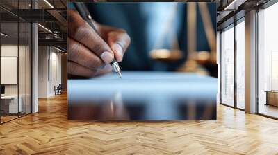 A close-up of a hand signing a document with a pen, the background features justice scales, symbolizing legal actions and authority in a professional office setting. Wall mural