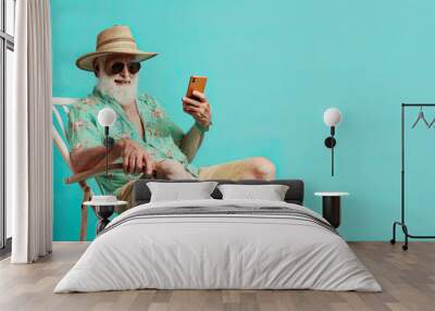 Mature male tourist sitting on a beach chair with a smartphone. an old man wearing beach clothes. Isolated on blue background Wall mural