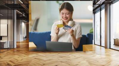 Smiling young asian woman holding credit card choosing bank payment method for online shopping on laptop at home Wall mural