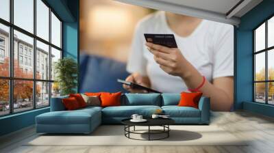 Woman is holding a credit card and using a smartphone for online shopping on a comfortable sofa at home Wall mural