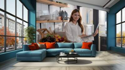 Young woman happily works on her laptop in a modern kitchen filled with fresh vegetables, embodying a healthy lifestyle Wall mural