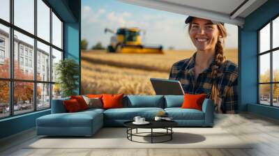 Young female farmer with digital tablet stands on wheat field on background with blurred yellow combine harvester. Wall mural