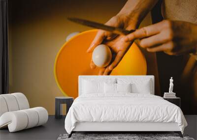 A woman is preparing to crack an egg using a knife, which she holds above a yellow bowl. This is a scene from a cooking process at home. Wall mural