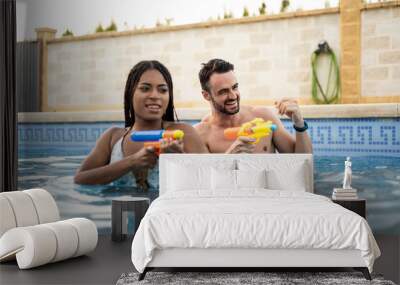 A couple of different ethnicities are playing in the pool with water guns. The young people are shooting with the guns while in the pool. Concept of couples having fun in a pool. Wall mural