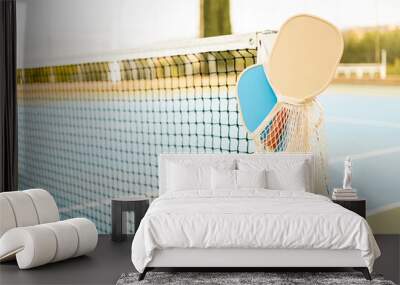 A mesh bag with a pair of pickleball rackets and two balls hooked to a net post on a sports court. Pickle ball concept.Sports with rackets Wall mural