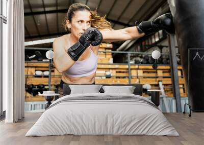 A middle-aged woman is training with a punching bag. The 50-year-old is hitting with her left arm with a boxing glove. Concept of female boxers between 40 and 50 years old. Boxing for adult women. Wall mural