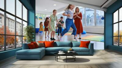 Small nursery school children with female teacher on floor indoors in classroom, doing exercise. Jumping over hula hoop circles track on the floor. Wall mural