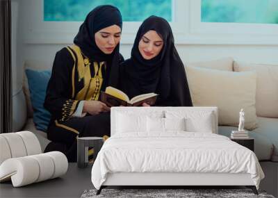 Two Young traditional Muslim women read Quran on the sofa before iftar dinner during a Ramadan feast at home Wall mural