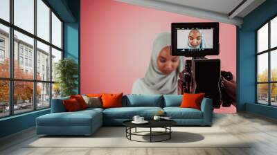 Videographer in digital studio recording video on professional camera by shooting female Muslim woman wearing hijab scarf plastic pink background. Wall mural