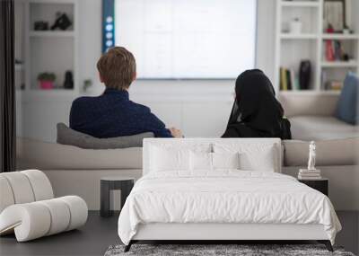 Young muslim couple woman wearing islamic hijab clothes sitting on sofa watching TV together during the month of Ramadan at modern home Wall mural