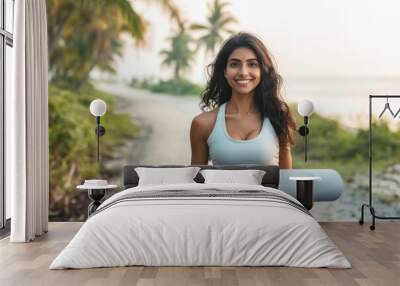 A beautiful Indian woman is going to do yoga in the morning by the seaside, carrying a yoga mat in her hands Wall mural