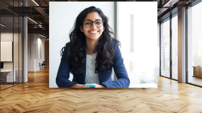 An Indian businesswoman in her 30s, wearing glasses, is happily smiling while sitting in her office in front of a window. Wall mural