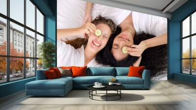 Two young attractive women in white bathrobes making facial mask holding slices of cucumber lying and chilling on bed posing to camera together in the bedroom, Women with beauty concept Wall mural