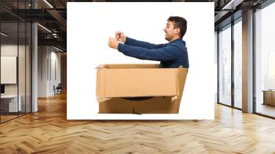 Full length side view of childish man sitting inside a cardboard box pretending to drive a new car isolated over white background. Joyful guy dreaming of buying a personal vehicle. Wall mural