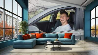 Joyful young man driving safe his new car, looking cheerful to camera. Contented teenage driver enjoying the ride, keeps hands on the steering wheel traveling, road trip concept Wall mural