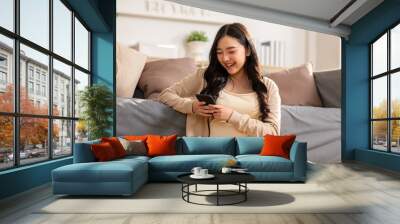 Happy asian young woman smile using smartphone surfing internet and enjoy with social media or online shopping at home. Modern wellness life of cute female looking at mobile phone fun and happiness Wall mural