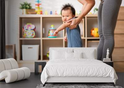 Mom helping and support baby learn to walk with love indoors at home. First steps of baby learning walk with family. Adorable asian baby pulling up to stand with mother support to develop leg muscle Wall mural