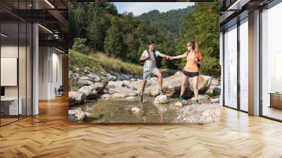 Active young hikers, man and woman walking over a mountain stream flowing among large beautiful rocks. Concept of nature exploring. Wall mural