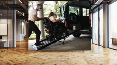 Assisting disabled woman on wheelchair with transport using accessible vehicle with ramp wearing a face mask. Wall mural