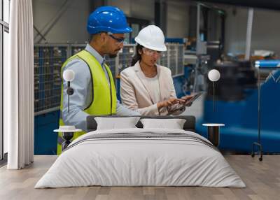 Female Indian factory supervisor is on-the-job training an African American male employee, in a blue helmet and yellow vest, to operate machinery. Wall mural