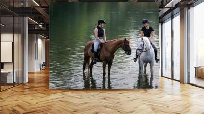 Riders, two young women riding beautiful horses down the calm river surrounded by the green grove Wall mural