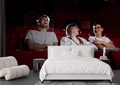 Three friends, a teenage girl and two boys smiling, watching a movie in an empty cinema with red chairs. Young adults and leisure concepts. Wall mural