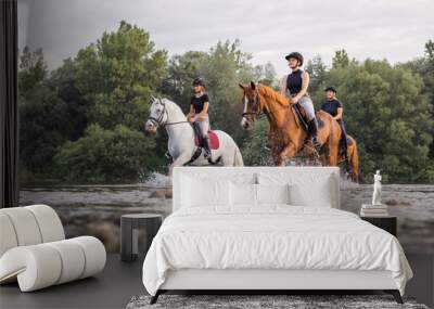 Three rider girls crossing the calm river water riding their beautiful horses on the cloudy summer day. Concept of equestrian leisure activity. Wall mural