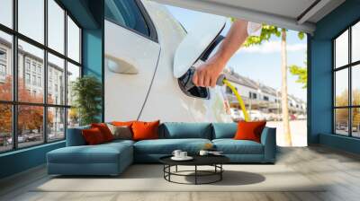 Woman plugging in the charger into a socket of her white electric car at a charging station in the street Wall mural