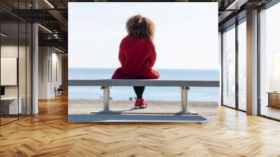 Rear view of a young curly woman wearing red denim jacket sitting on a bench while looking away to horizon over sea Wall mural