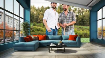 Two men in plaid shirt standing in oat field with forest in background and talking Wall mural