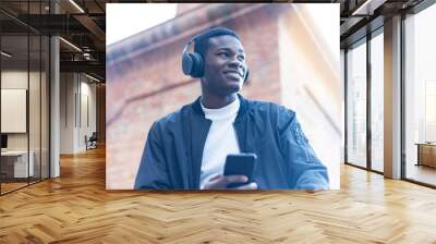 Young Black man standing in the street while listening music by headphones Wall mural