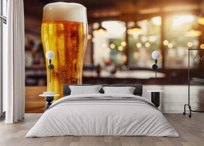 A pint of beer rests on a wooden bar table, the amber liquid glistening under the bar lights. The beer glass is surrounded by other barware and drinkware Wall mural