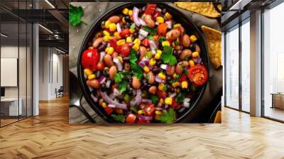 Vertical view of a black bean salad featuring black-eyed peas, red peppers, onions, corn, cherry tomatoes, and cilantro, presented in a dark bowl alongside tortilla chips and nachos on a surface. Wall mural