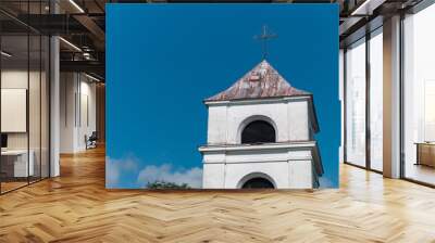 Close-up view of a white church bell tower against a bright blue sky. The tower is adorned with a cross at the top, symbolizing its religious purpose. The tower's white walls are slightly weathered. Wall mural