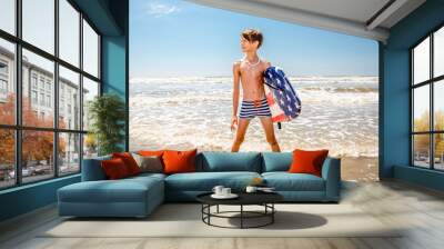 Young boy in striped swim trunks standing in the water at the beach with his boogie board Wall mural