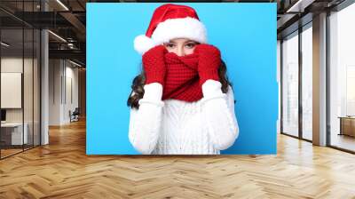 Beautiful girl in santa hat, gloves and scarf on blue background Wall mural