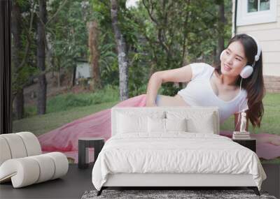 Young asian woman sitting in fresh spring grass listening to music and smiling with pleasure. Beautiful female expecting child having rest outside in front of her home Wall mural