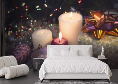 A festive still life with a purple and two white candles of different size, a light patterned gift box with a dark shiny bow, a lilac pinecone and a ball. Dark Christmas background. Blurred bokeh Wall mural