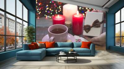 Hand holding a white gift box with a brown bow, a pink porcelian cup of tea, a saucer, two red and one white burning candles and fairylights on the back, a colored wooden background Wall mural